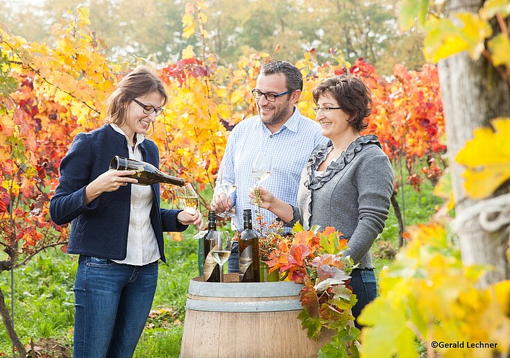 Monika Neustifter schenkt ein Glas Wein ein. Zwei Gäste halten ein Glas Wein in der Hand. Alle stehen im Weingarten der schon bunt vom Herbst ist. In der Mitte steht ein Weinfass mit Weinflaschen.