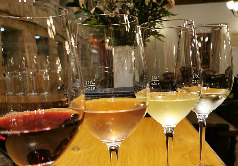 Vier Weingläser stehen nebeneinander farblich gereiht, beginnend mit rot, rosé, trübes weiß, weiß.