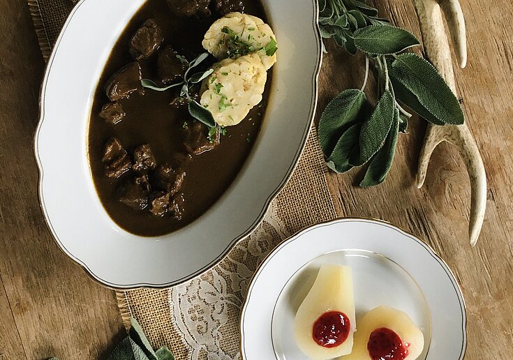 In einem weißen, ovalen Teller ist ein Wildragout mit Knödel angerichtet. Daneben ist ein weiteres Teller mit zwei Birnenhälften mit Preiselbeeren in der Mitte. Auf dem Tisch befindet sich auch ein Hirschgeweih als Dekoration.