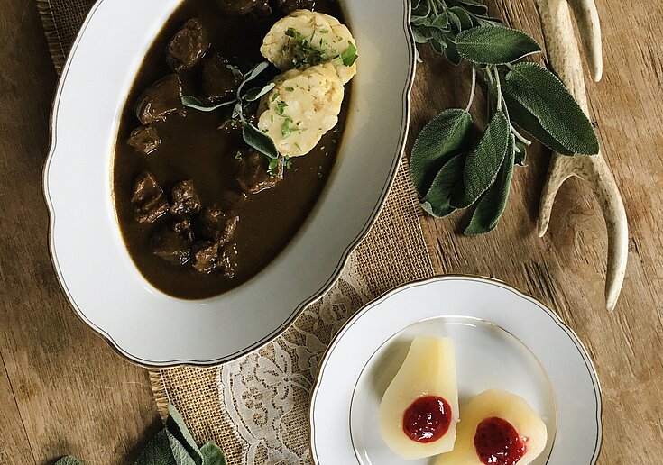 In einem weißen, ovalen Teller ist ein Wildragout mit Knödel angerichtet. Daneben ist ein weiteres Teller mit zwei Birnenhälften mit Preiselbeeren in der Mitte. Auf dem Tisch befindet sich auch ein Hirschgeweih als Dekoration.