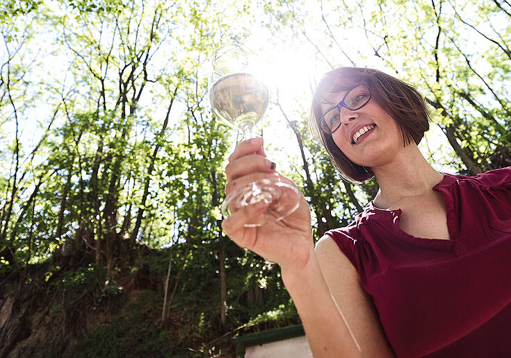 Monika Neustifter wird von unten fotografiert. Sie hält ein Weinglas in der Hand. Ober ihr sieht man Baumspitzen und Lichtstrahlen.
