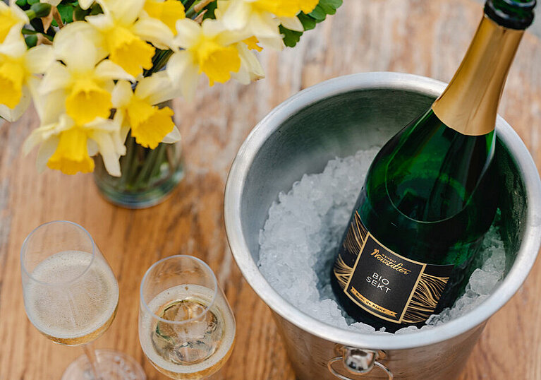 A bottle of BIO Blanc de Blancs sparkling wine. On a table stands a champagne cooler with a bottle of Neustifter BIO Blanc de Blancs sparkling wine, two glasses and a vase of yellow flowers.