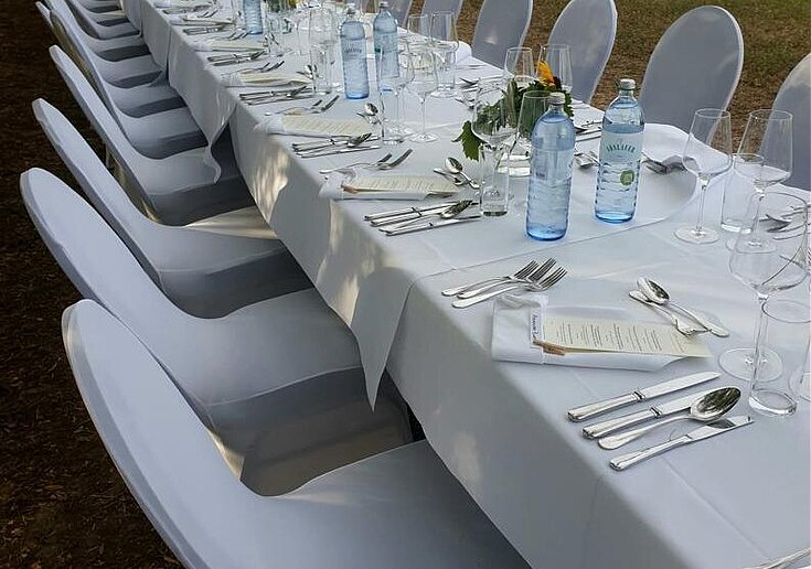 Eine lange weiße, geschmückte Tafel mit weißen Sesseln stehen in der Natur. Dahinter sieht man Weingärten. Die lange Tafel ist eingedeckt mit Servietten, Gläser und Besteck.