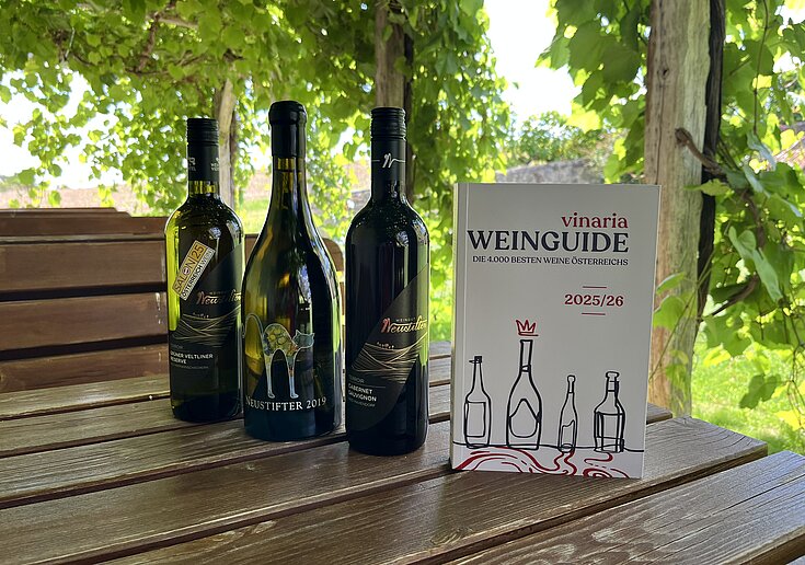 Das Vinaria Magazin und 3 Flaschen Neustifter Wein stehen auf einem Tisch unter der Laube