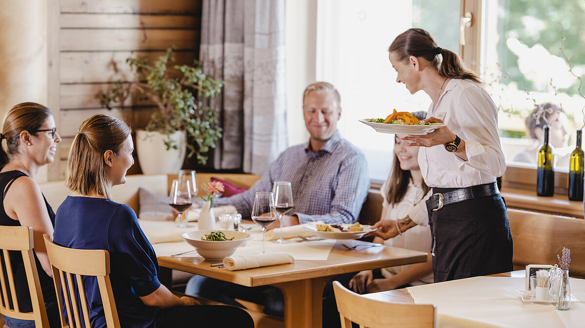 Eine Servicemitarbeiterin serviert einer Familie leckere Gerichte in der Gaststube vom Wein.Restaurant Neustifter.