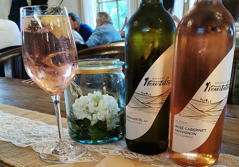 Eine Flasche BIO Rosé Cabernet Sauvignon und eine Flasche BIO Weinviertel DAC Grüner Veltliner stehen am Tisch mit einem Glas Wein und zwei Speisekarten des offenen Kellers.