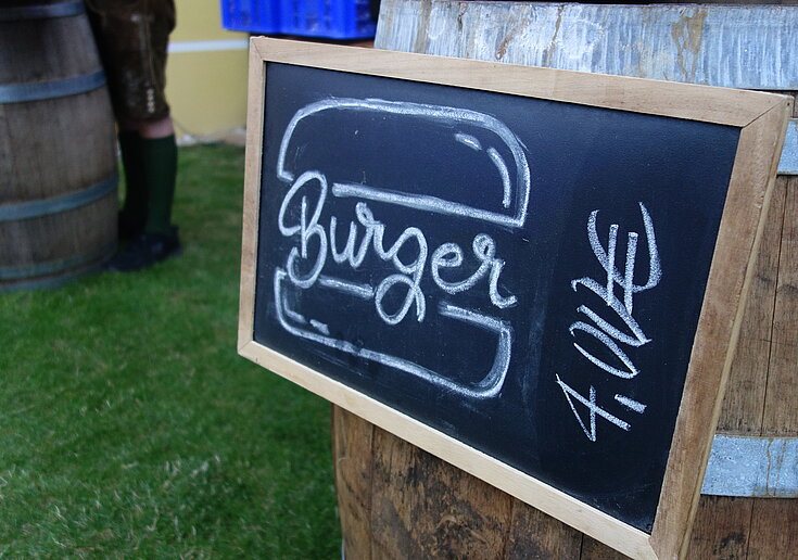 Auf einer schwarzen Tafel steht in weißer Schrift "Burger". Die Tafel hängt auf einem Weinfass und steht im Garten.