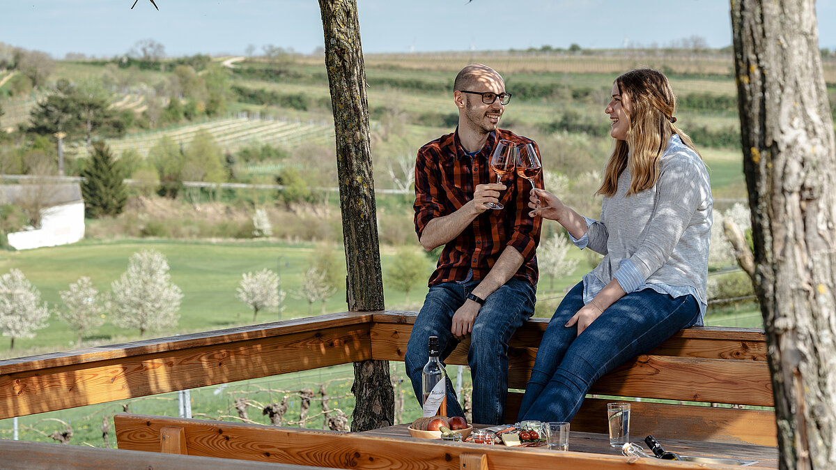 Ein Pärchen sitzt auf dem Geländer der Sonnenterrasse und stoßt mit einem Glas Wein an. Im Hintergrund hat man eine schöne Aussicht auf den Weingarten.