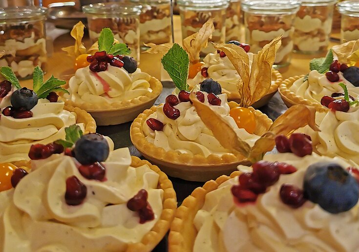 In kleinen Schüsselchen auf Mürbteig ist eine weiße Vanillecreme mit verschiedenen Früchten oben drauf. Die Törtchen stehen auf einem Buffet.