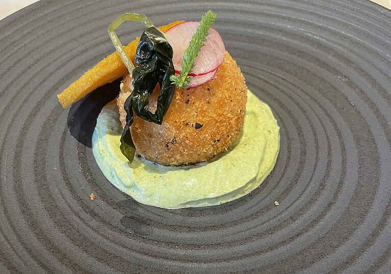 Veganes Menü Ein frittiertes Gemüsebällchen auf Wasabicreme liegt auf einem schwarzen, runden Teller.