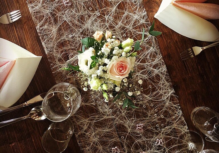 Muttertagsbuffet gedeckter Tisch In einer Vase sind geschnittene Rosen mit grünem Schleierkraut. Unter der Vase ist ein weißer Seidentischläufer. Zwei gefaltete Servietten, sowie Gabel, Messer und Gläser sind auf dem Tisch eingedeckt.