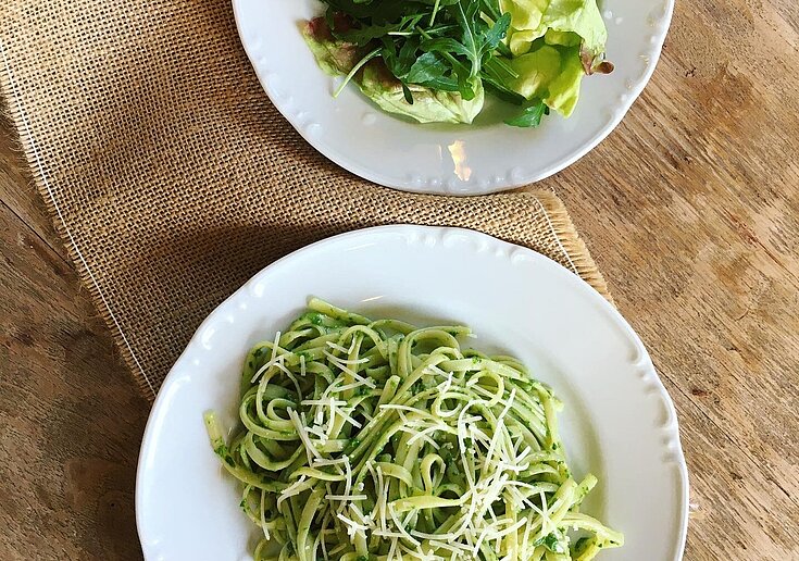 Auf dem rustikal gedeckten Tisch, sind zwei weiße runde Teller. Auf dem vorderen Teller wurden Spaghetti mit Bärlauchpesto angerichtet. Auf dem hinteren Teller ist ein grüner Salat angerichtet.