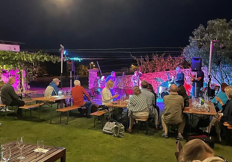 Der Gastgarten vom TOP Heuriger Wein.Küche ist voll mit Tischen und Gästen. Es ist Nacht. Im Hintergrund steht eine Jazzband und spielt.