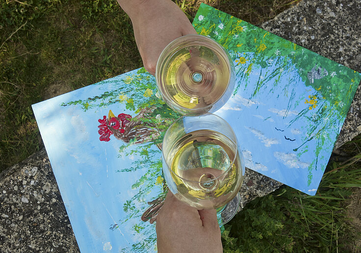 Drink x Create draußen Zwei Landschaftsbilder im Hintergrund, im Vordergrund zwei Wiengläser, die anstoßen.