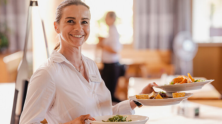 Eine Kellnerin hält drei Teller in der Hand und lächelt in die Kamera.