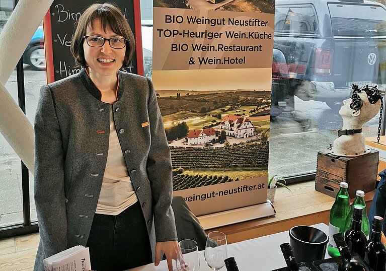 Monika Neustifer am Weintour Opening Monika Neustifter steht hinter dem Präsentiertisch beim Weintour Opening. Hinter ihr steht ein Rollup mit Bildern vom Wein.Gut und Wein.Hotel Neustifter.