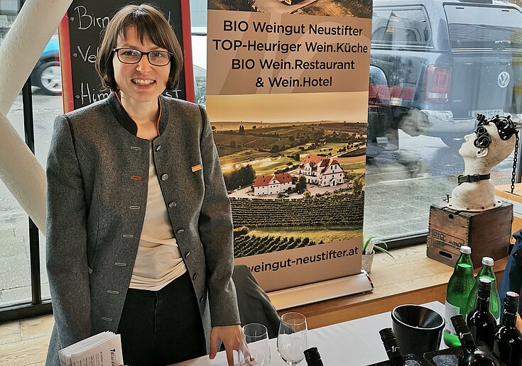 Monika Neustifter steht hinter dem Präsentiertisch beim Weintour Opening. Hinter ihr steht ein Rollup mit Bildern vom Wein.Gut und Wein.Hotel Neustifter.