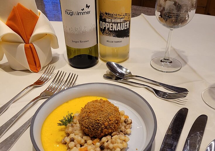 Auf einem gedeckten Tisch wurde ein Teller Ganslkroketten mit Kürbiscappuccino serviert. Daneben steht eine Weinflasche vom Weingut Hugl-Wimmer und eine Flasche vom BIO Weingut Valentin Oppenauer.