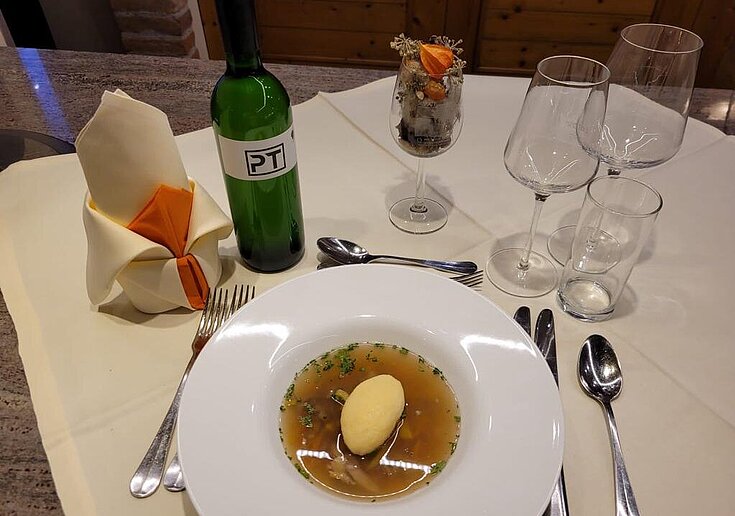 Auf einem gedeckten Tisch wurde ein Teller mit Ganslsuppe und Grießnockerl serviert. Daneben steht eine Flasche Wein vom Weingut Peter Terscinar.