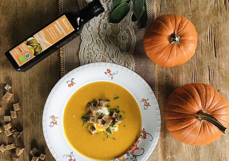 Auf dem rustikal geschmückten Tisch, steht in der Mitte ein Suppenteller mit Kürbiscremesuppe. In der Kürbiscremesuppe befinden sich Weißbrotcroutons. Weiters sieht man auf dem Tisch zwei orange Kürbisse und eine Flasche mit BIO Kürbiskernöl vom Weingut Neustifter.