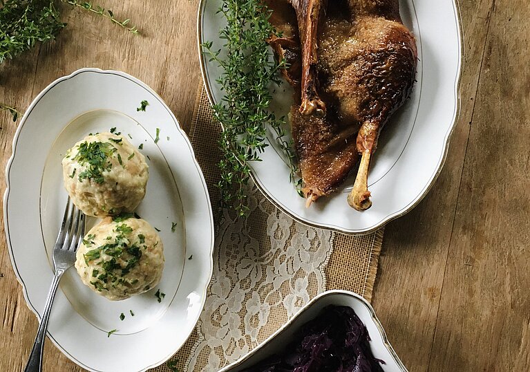 Ein Teller mit genussvolle Ganslkeulen, zwei Kartoffelknödel und eine Schüssel mit selbstgemachtem Rotkraut stehen auf einem Holztisch.