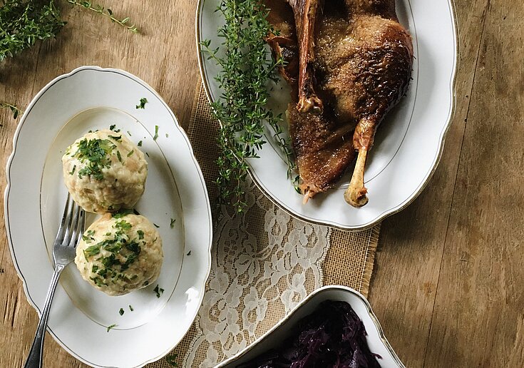 Ein Teller mit genussvolle Ganslkeulen, zwei Kartoffelknödel und eine Schüssel mit selbstgemachtem Rotkraut stehen auf einem Holztisch.