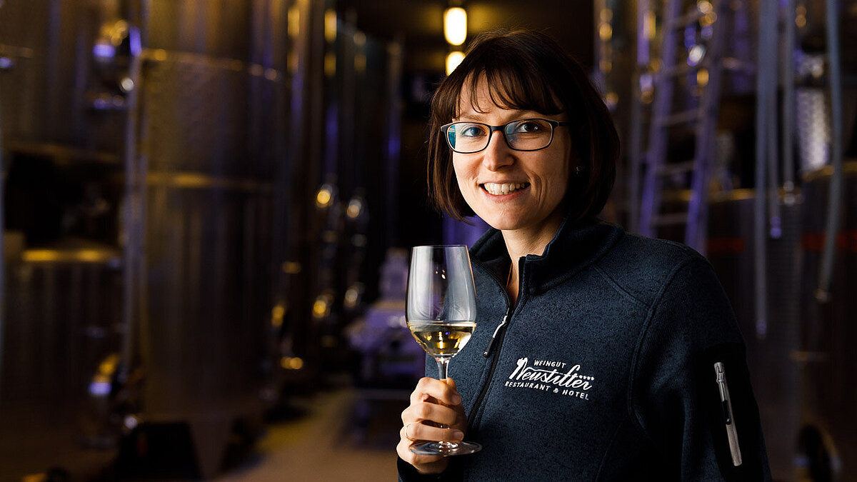 Monika Neustifter steht im Weinkeller vor mehreren Stahltanks. Sie hält ein Weißweinglas in der Hand und lächelt.