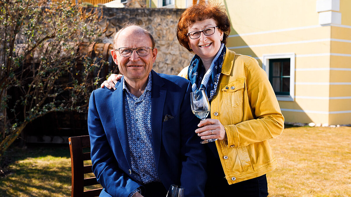 Karl Neustifter sitzt auf einem Hocker und Brigitte Neustifter steht bei ihm. Sie haben beide ein Glas Wein in der Hand.