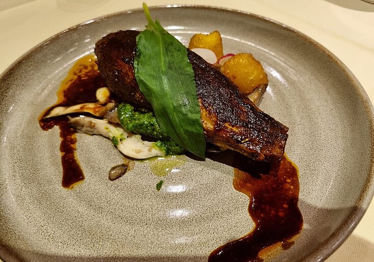 Veganes Menü gegrillte Melanzani Das runde Teller ist grau gesprenkelt. Auf dem Teller ist eine ganze gegrillte Melanzani, die mit Gemüse gefüllt ist. Bratkartoffeln sind die Beilage.