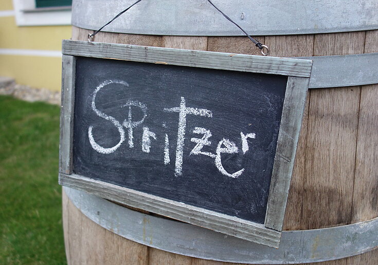 Auf einer schwarzen Tafel steht in weißer Schrift "Spritzer". Die Tafel hängt auf einem Weinfass und steht im Garten.