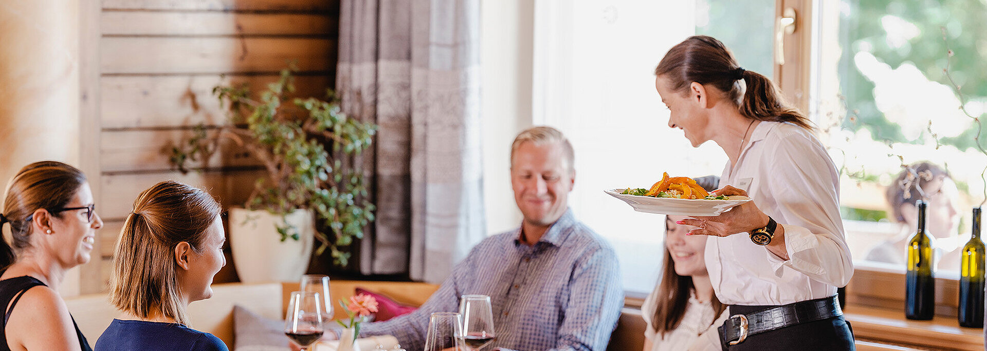 Eine Servicemitarbeiterin serviert einer Familie leckere Gerichte in der Gaststube vom Wein.Restaurant Neustifter.