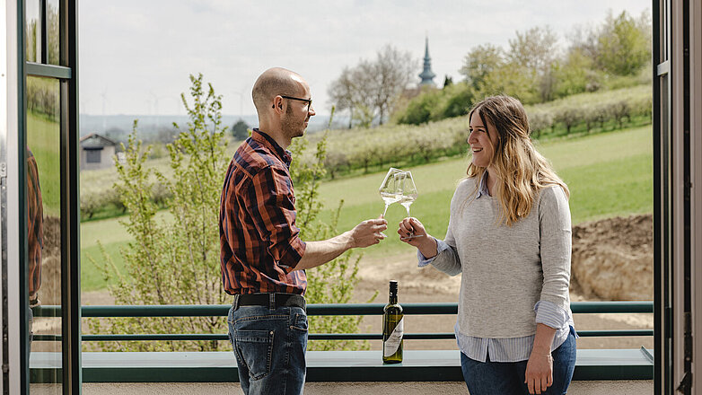 Ein Pärchen steht auf dem Balkon des Wein.Hotel Neustifters und prostet sich zu. Auf dem Geländer steht eine Flasche Neustifter Wein. Im Hintergrund kann man die Stadtpfarrkirche Poysdorf erkennen.