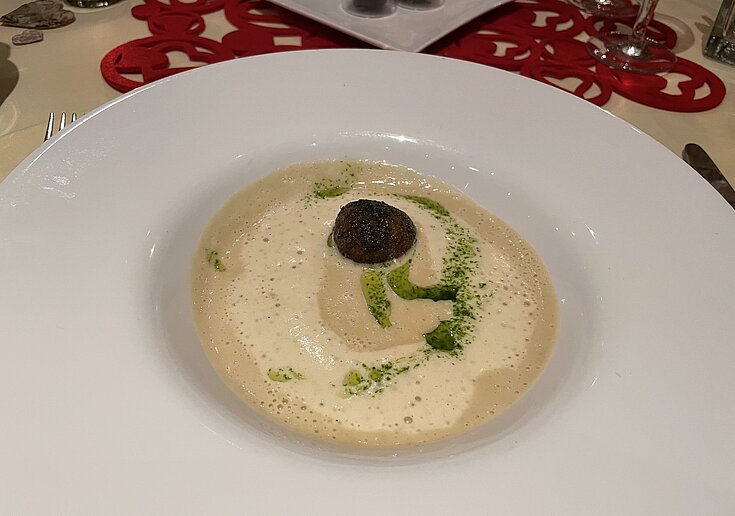 Valentinstagsmenü Veltlinercremesuppe In dem weißen Suppenteller mit breitem Rand ist eine weiße Cremesuppe mit Schlagobershaube.