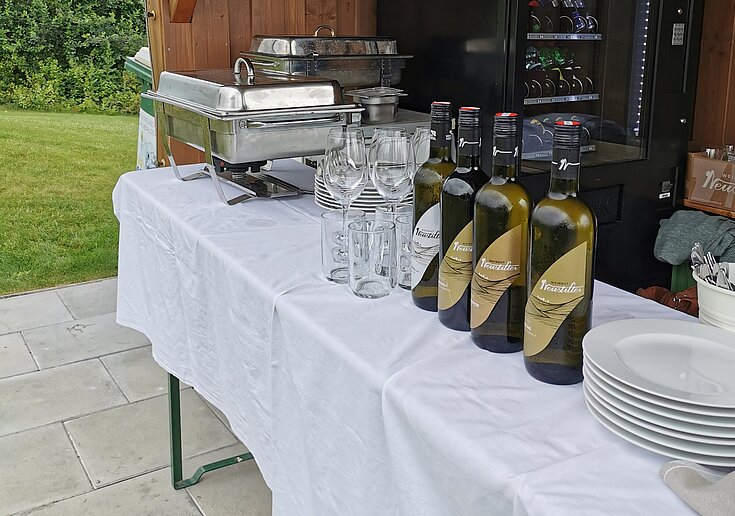Für die Halfwayverpflegung des Weincup Golfturniers gibt es ein Buffet mit weißen Tischtuch. Darauf stehen Weine, Gläser, Teller und Chevingtische mit warmen Essen.