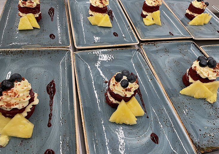 Valentinstagsmenü Schokotörtchen Mehrere eckige graue Teller stehen nebeneinander. Auf den Teller sind Schokotörtchen mit weißer Creme und Heidelbeeren oben drauf. Ananasecken verzieren das Teller.