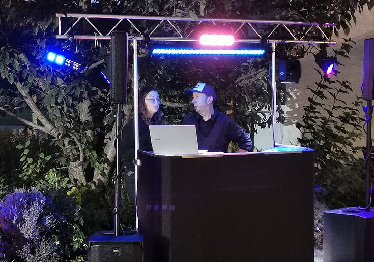 Hinter dem schwarzen DJ-Pult steht DJ Illy und ein Gast. Es ist Nacht und bunte Lichter flackern.