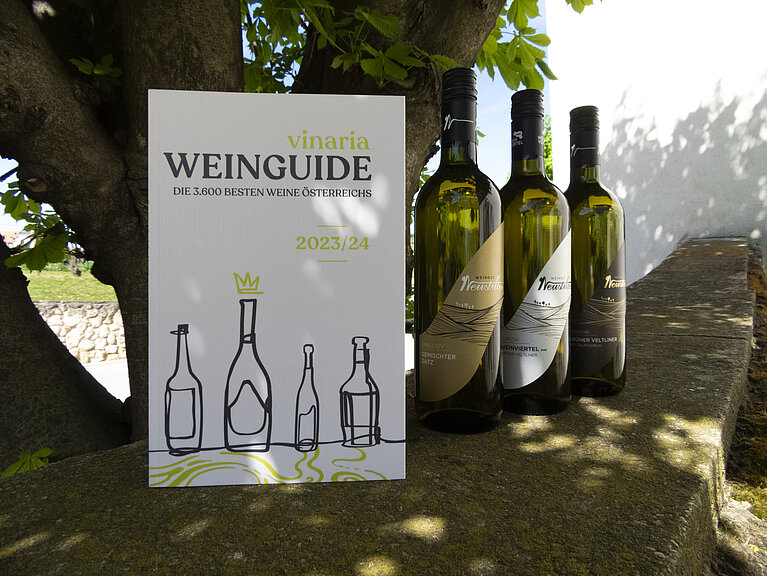 Vinaria 2023/2024 Das Vinaria Magazin und drei Flaschen Neustifter Wein stehen auf einer Mauer unter einem Baum.