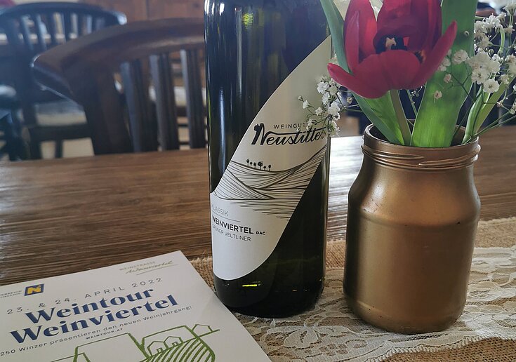 Auf dem Tisch steht eine Flasche Neustifter Wein und daneben eine braune Vase mit einer lila Tulpe mit grünem Schleierkraut. Davor liegt ein Werbeheft der Weintour.