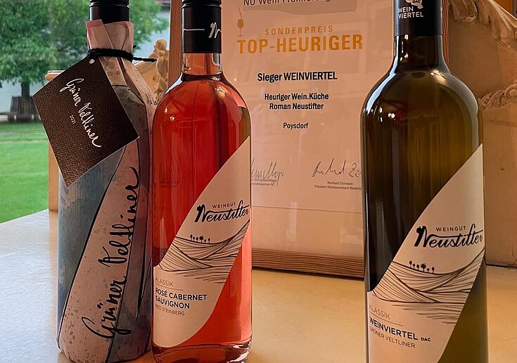 Eine Flasche BIO Grüner Veltliner unfiltriert 2021, eine Flasche BIO Rosé Cabernet Sauvignon 2023 und eine Flasche BIO Weinviertel DAC Grüner Veltliner 2023 stehen vor der Urkunde des TOP Heuriger Sonderpreises der Wein.Küche Neustifter.