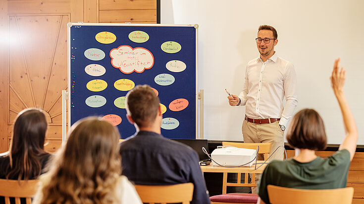 Ein Vortragender hält ein Seminar ab. Neben ihm steht eine Pinnwand.