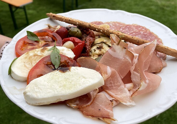 Auf einem weißen, runden Teller liegen Weinviertler Anti-Pasti geschmackvoll angerichtet. Dahinter erkennt man den Gastgarten der TOP Heuriger Wein.Küche.