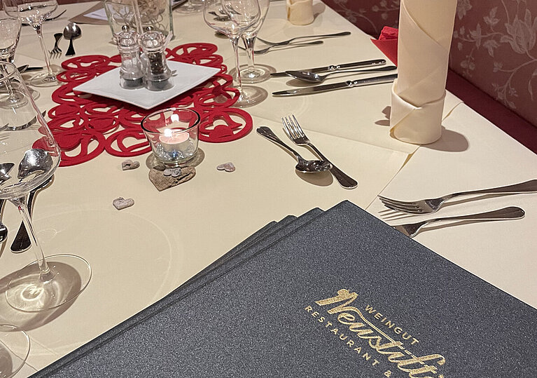 Valentinstagsmenü Romantisches Gedeck Man sieht einen romantisch geschmückten Tisch mit Servietten und Kerzen. Am Tisch liegt die Speisekarte vom Wein.Hotel Neustifter.