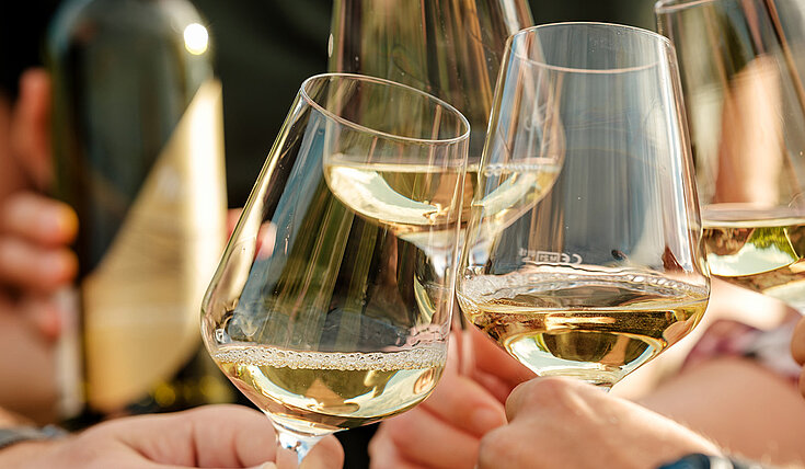 Gläser mit Weißweinen stoßen zusammen. Im Hintergrund sieht man eine Flasche Neustifter Wein aus der Exklusiv Linie.