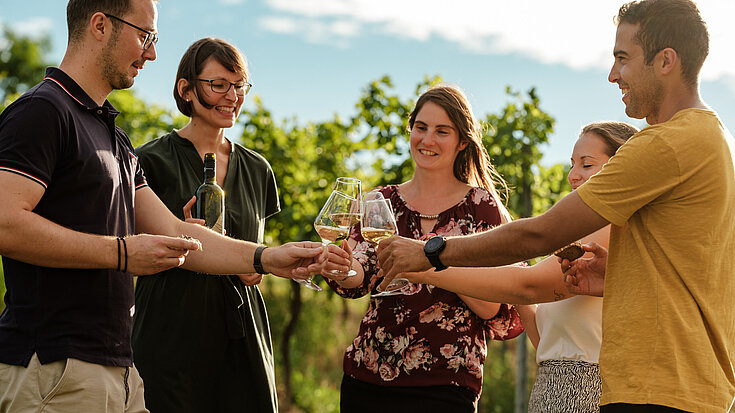 Eine Gruppe junger Menschen steht im Weingarten mit Monika Neustifter und verkostet eine Flasche Wein.