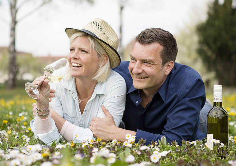 Ein Pärchen liegt in einer Blumenwiese mit einem Glas Wein in der Hand und lacht.