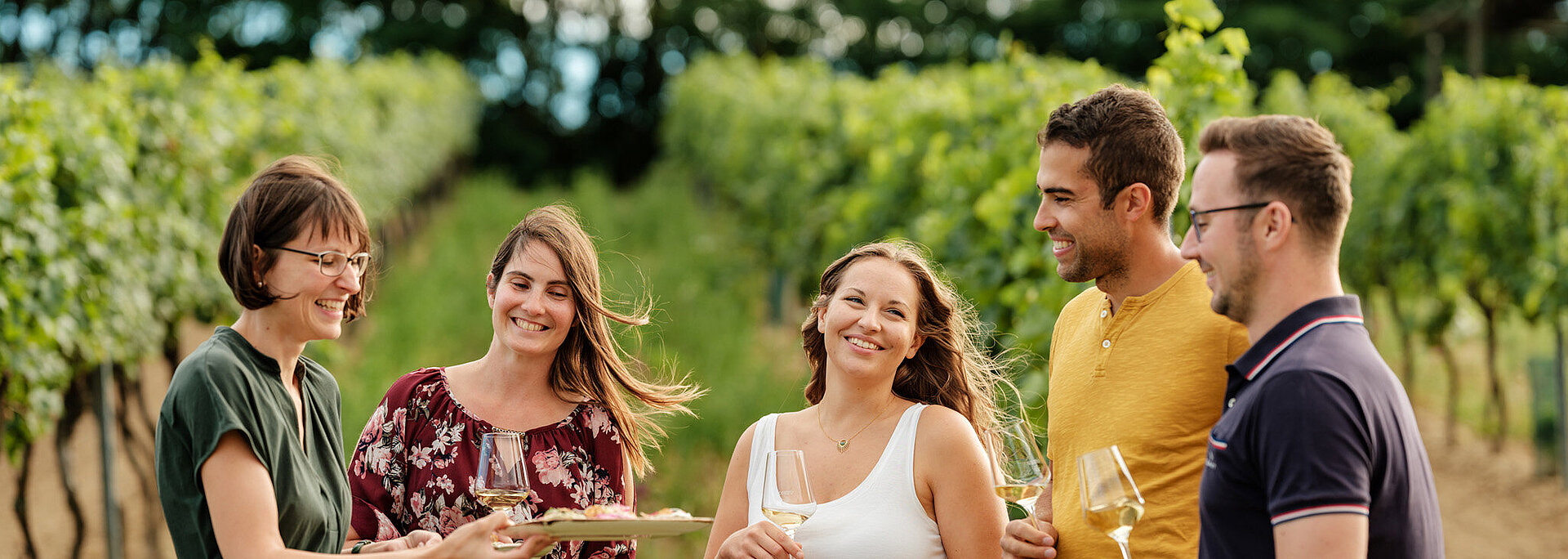 Eine Gruppe junger Menschen steht mit Monika Neustifter im Weingarten. Monika Neustifter bietet der Gruppe Brötchen und ein Glas Wein an.