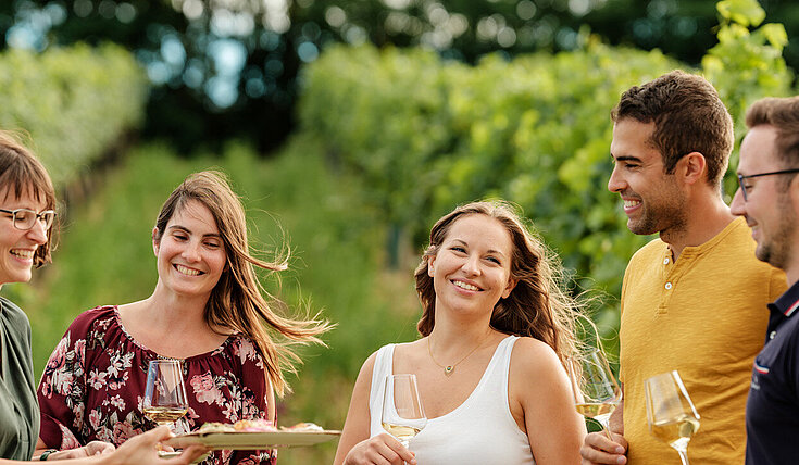 Eine Gruppe junger Menschen steht mit Monika Neustifter im Weingarten. Monika Neustifter bietet der Gruppe Brötchen und ein Glas Wein an.