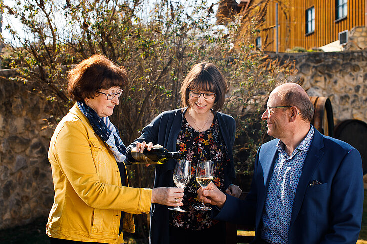 Brigitte, Monika und Karl Neustifter stehen zusammen. Monika Neustifter schenkt ihren Eltern ein Glas Wein ein.