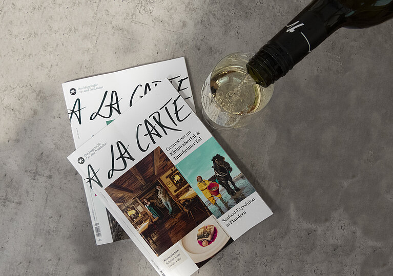 Zwei A la Carte Magazine liegen am Tisch und es wird ein Glas Weißwein eingeschenkt.