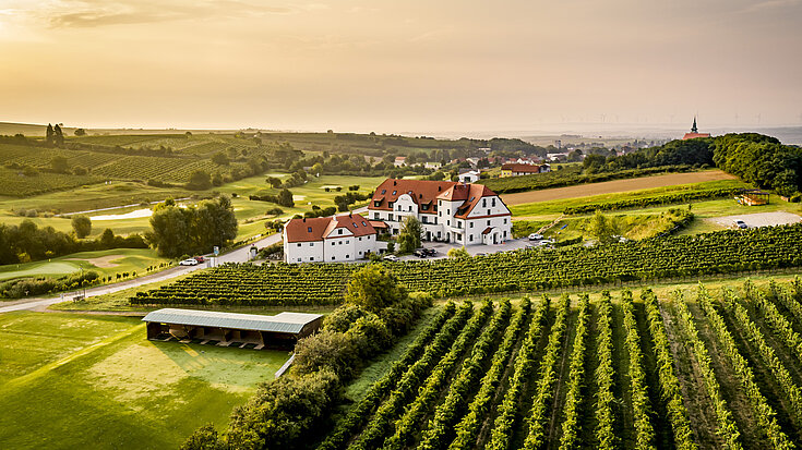 Eine Drohnenaufnahme vom Wein.Hotel Neustifter. Im Vordergrund stehen die Weingärten und die Driving Range des Golfplatzes nebenan.